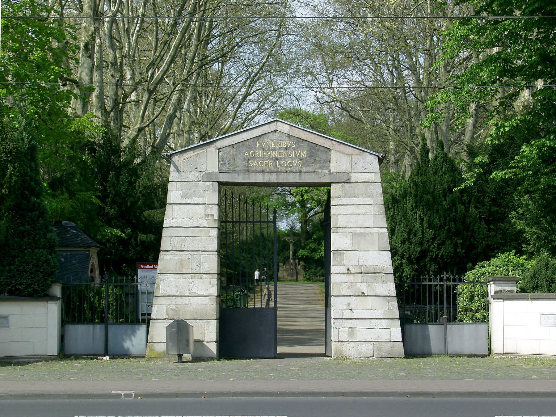 Frontansicht des steinernen Torbaus am Eingang Aachener Straße mit geöffnetem Gittertor und goldener lateinischer Inschrift auf Melaten-Friedhof