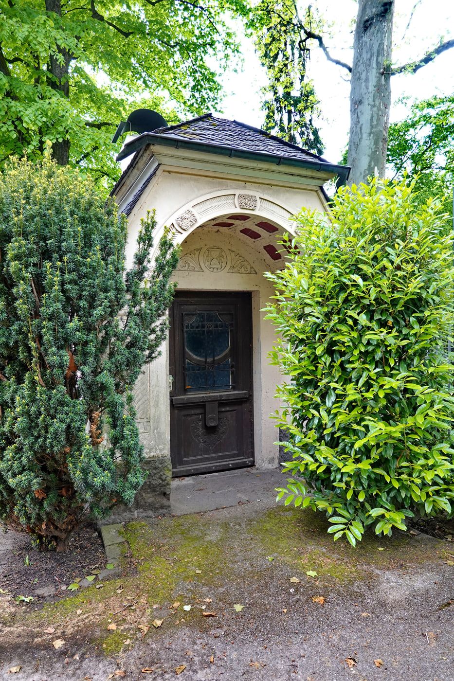 Kleines helles Pförtnerhäuschen mit Rundbogenportal, dekoriertem Türfeld und dunklem Schieferdach zwischen hohen Sträuchern auf Melaten-Friedhof