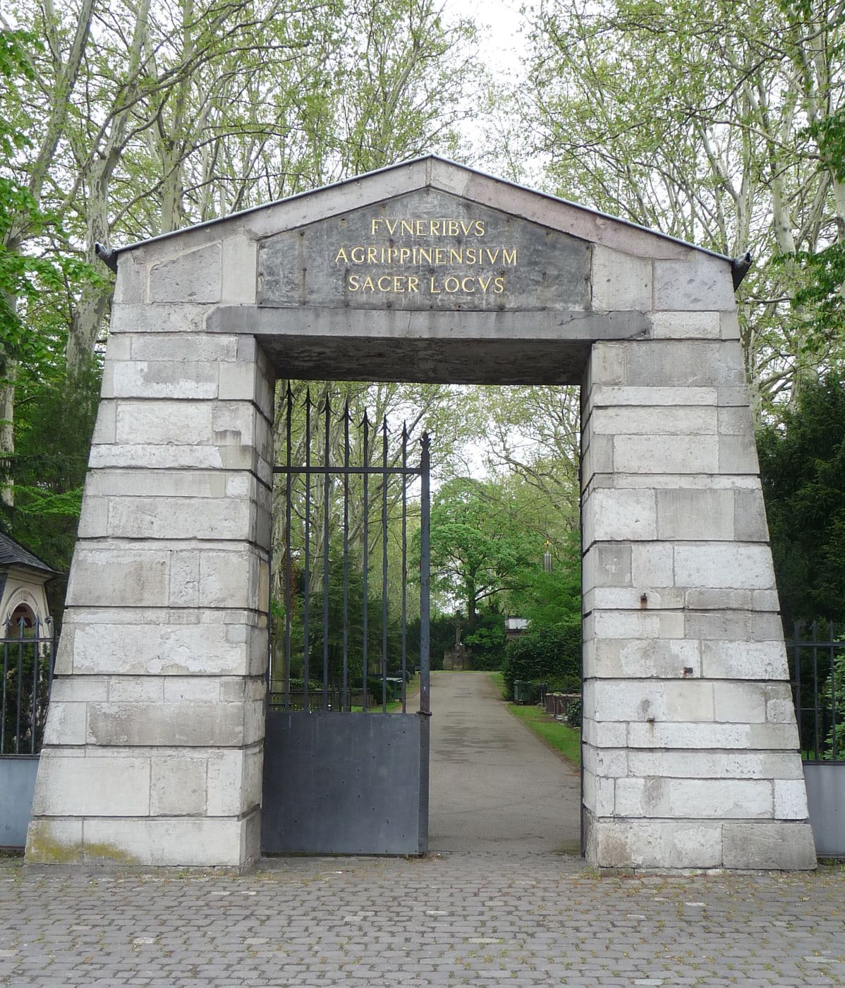 Nahansicht des steinernen Torbaus an der Aachener Straße mit geöffnetem Spitzengitter und goldener lateinischer Inschrift auf Melaten-Friedhof